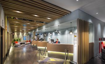 a modern hotel lobby with a reception desk , where two people are standing and a sign indicating the name of the establishment at Ibis Melbourne Hotel and Apartments