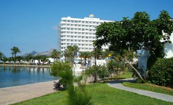 a large white building with a hotel in the background , surrounded by a park - like setting at Club Mac Alcudia