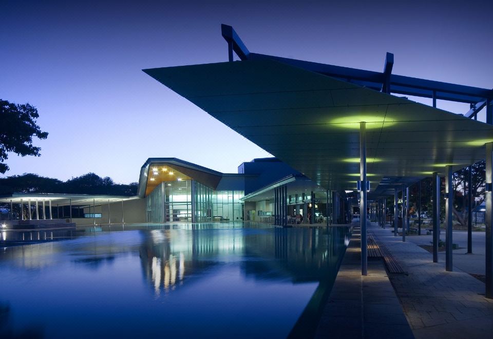 a modern building with a large glass roof , reflecting in a pool at night , surrounded by people at Jacana Apartments