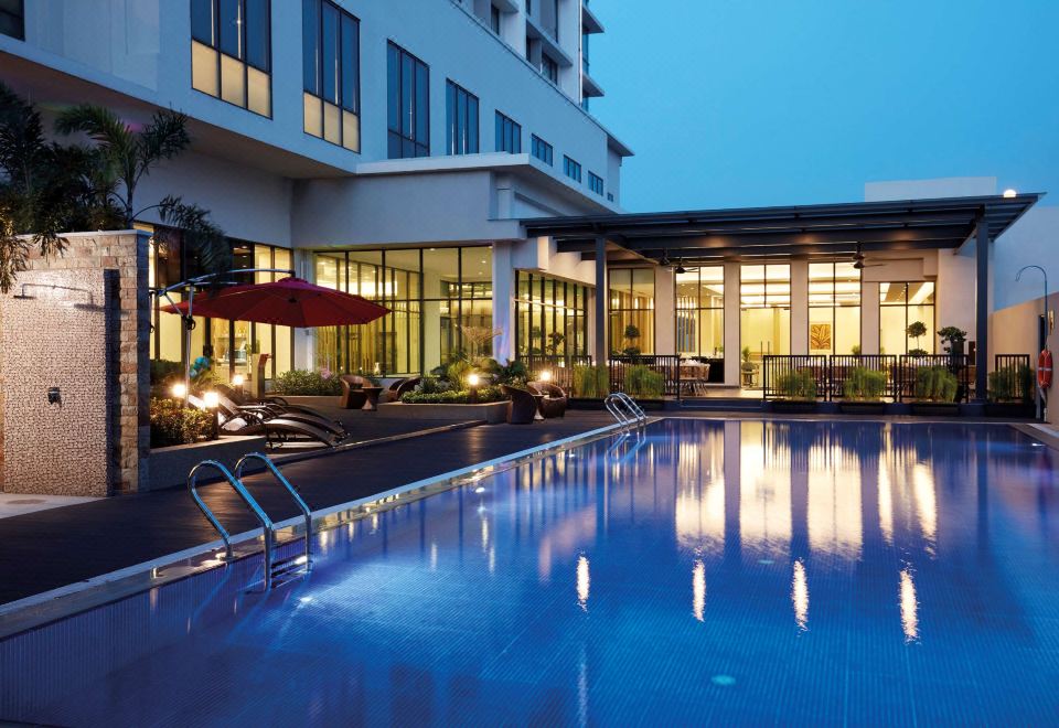 an outdoor swimming pool surrounded by a hotel building , with lounge chairs and umbrellas placed around the pool at Hilton Garden Inn Puchong