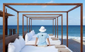 a person wearing a blue shirt is standing on a bed with white pillows and blankets , overlooking the ocean at White Pearl Resorts
