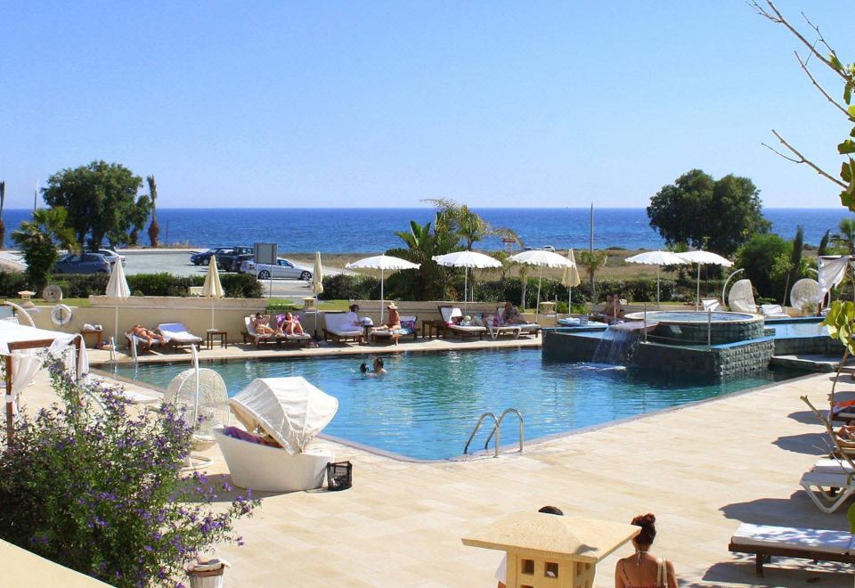 a swimming pool with umbrellas and people , overlooking the ocean , under a clear blue sky at E-Hotel Larnaca Resort & Spa