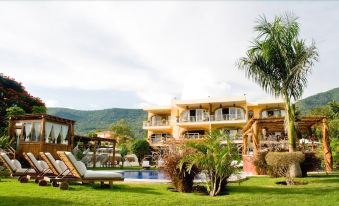 a large building with balconies is surrounded by a pool and palm trees , with mountains in the background at El Chante Spa Hotel