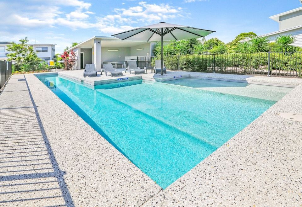 a large , empty swimming pool with a blue water surface and white tiles surrounding it , under an open - air structure with umbrellas at Direct Collective - Breeze on Brightwater