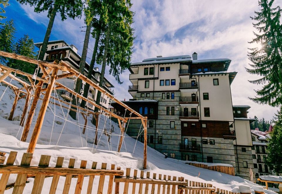 a snow - covered hill with a wooden walkway and several buildings , including a hotel in the background at Green Life Family Apartments Pamporovo
