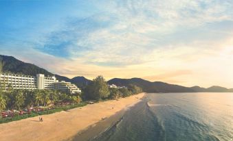 a beach with a few people in the water and mountains in the background during sunset at PARKROYAL Penang Resort