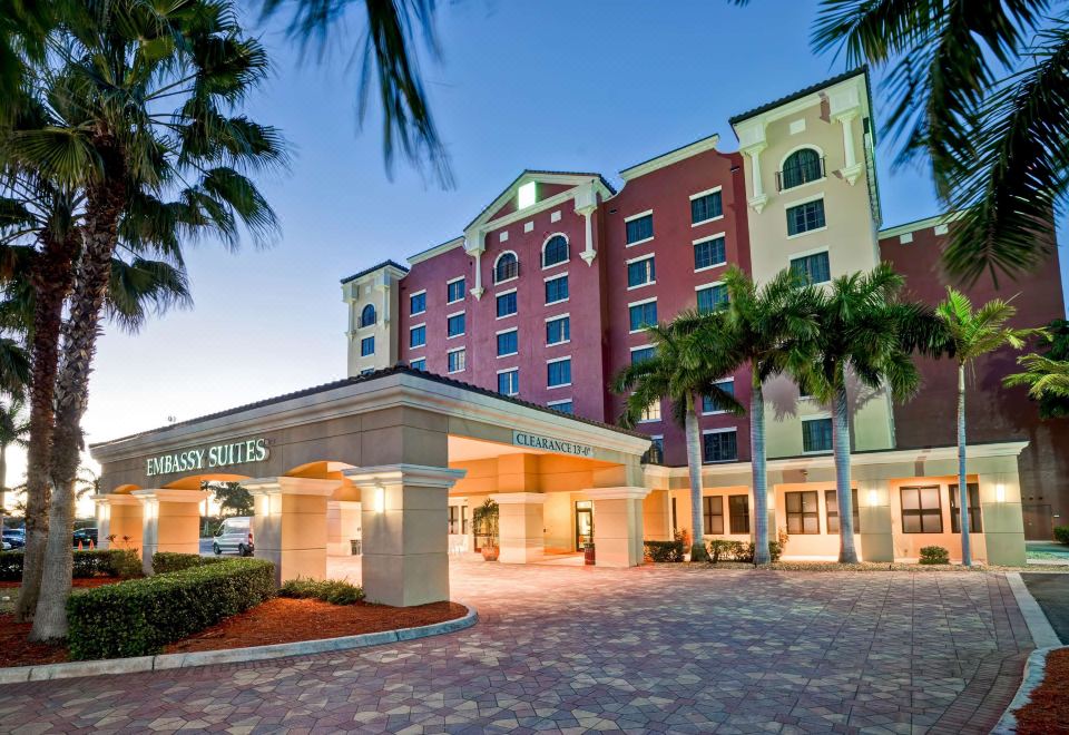 a large hotel with a red and white exterior , surrounded by palm trees and lit up at night at Embassy Suites by Hilton Fort Myers Estero