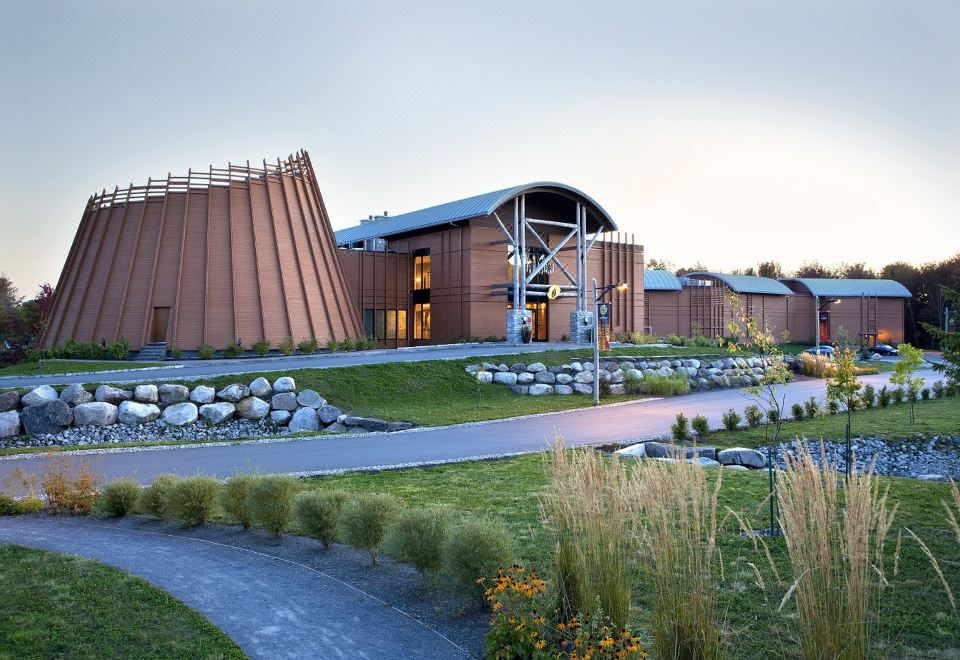 a modern building with a large wooden structure , surrounded by lush greenery and flowers , under a cloudy sky at Hotel-Musee Premieres Nations