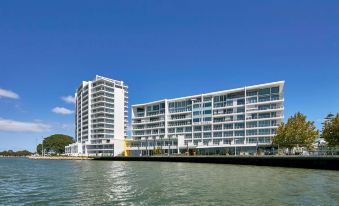a large white building with multiple floors , situated near the water and surrounded by trees and other buildings at The Sebel Mandurah