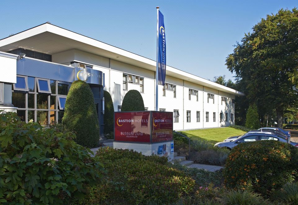 a large white building with a blue banner in front of it , surrounded by grass and trees at Bastion Hotel Bussum Hilversum