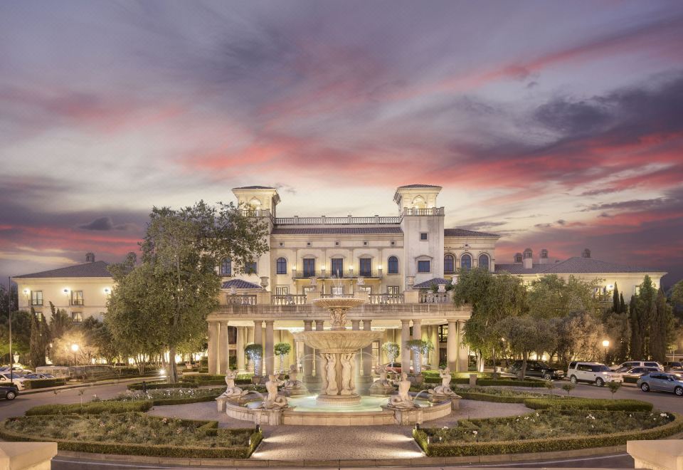 a large white building with columns is surrounded by a garden and trees , with a fountain in front of it at Palazzo Hotel