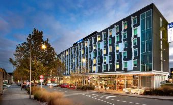 a modern , multi - story building with black and blue exterior facades , surrounded by trees and buildings , and lit at night at Ibis Budget Auckland Airport