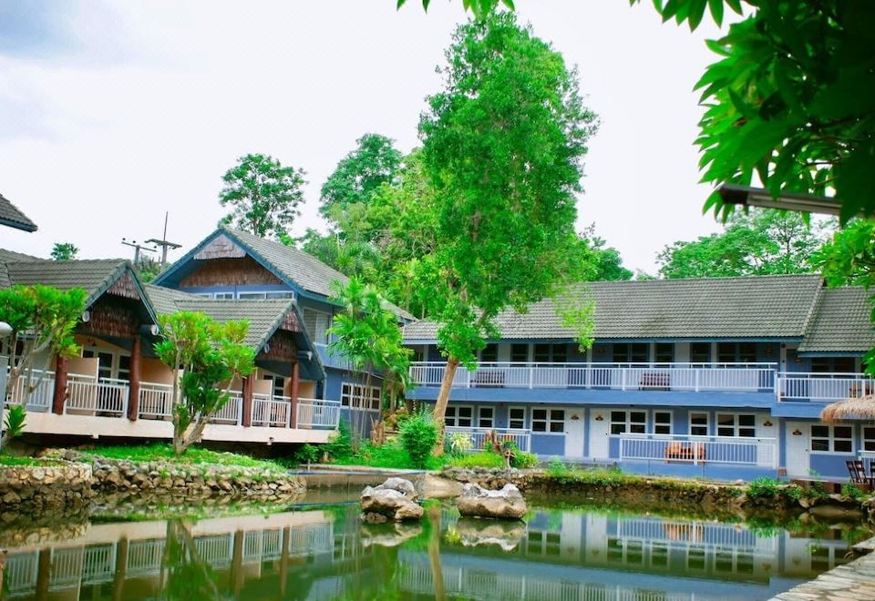 a large blue building surrounded by a pond , with trees and a small body of water nearby at Baanrai Riverkkwai Resort