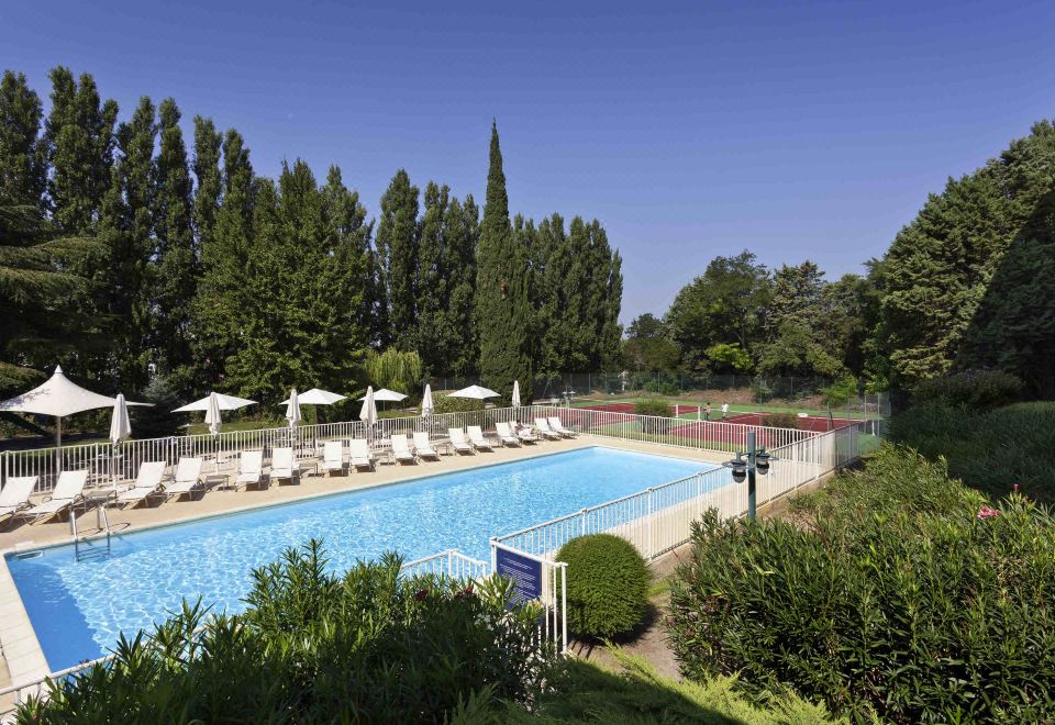 a large outdoor swimming pool surrounded by lush greenery , with several lounge chairs and umbrellas placed around the pool at Novotel Avignon Nord