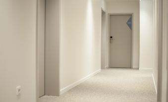 a long , empty hallway with white walls and a gray door at the end , leading to a bathroom at Bayerischer Hof