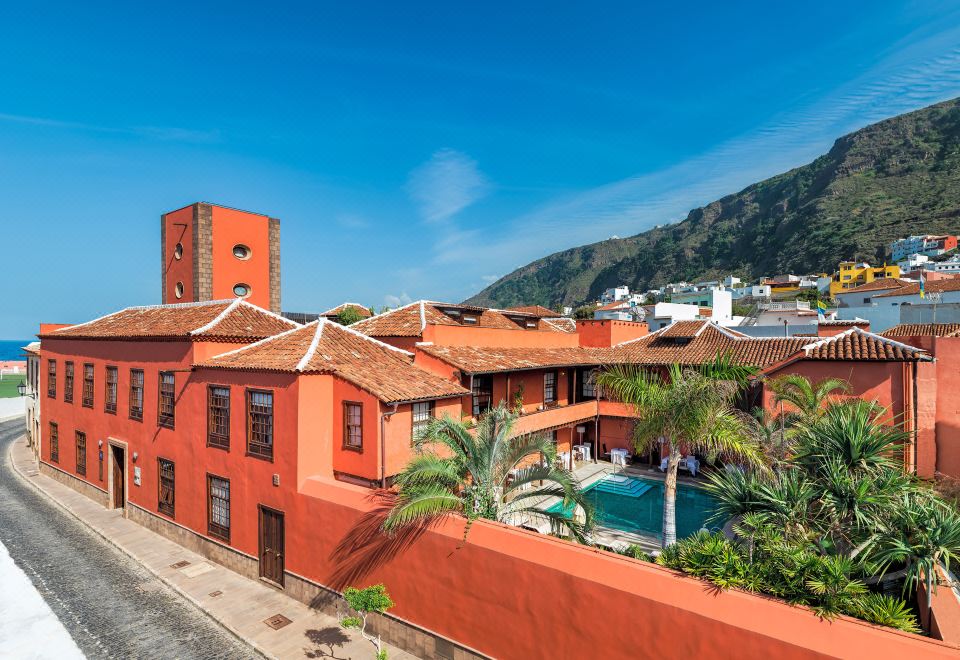 a red building with a pool and palm trees in front of it , surrounded by mountains at Hotel San Roque