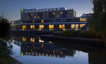 a large hotel building situated on the edge of a body of water , with lights reflecting in the water at Holiday Inn London - Brentford Lock