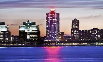 a city skyline at night , with tall buildings illuminated by colorful lights reflecting on the water at W Hoboken