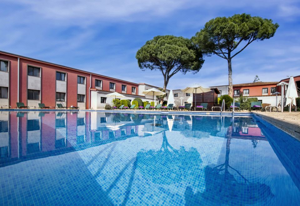 a large swimming pool is surrounded by a brick building and trees , with a blue sky above at Salles Hotel Aeroport de Girona