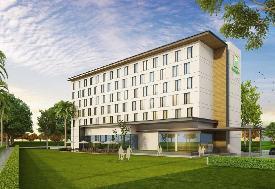 a large white building with many windows is surrounded by green grass and trees , with a person walking in the foreground at Holiday Inn Express Kolkata Airport