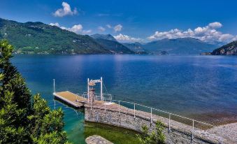 a serene lake with a pier extending into the water , surrounded by lush greenery and mountains in the background at Villa Lario Resort Mandello
