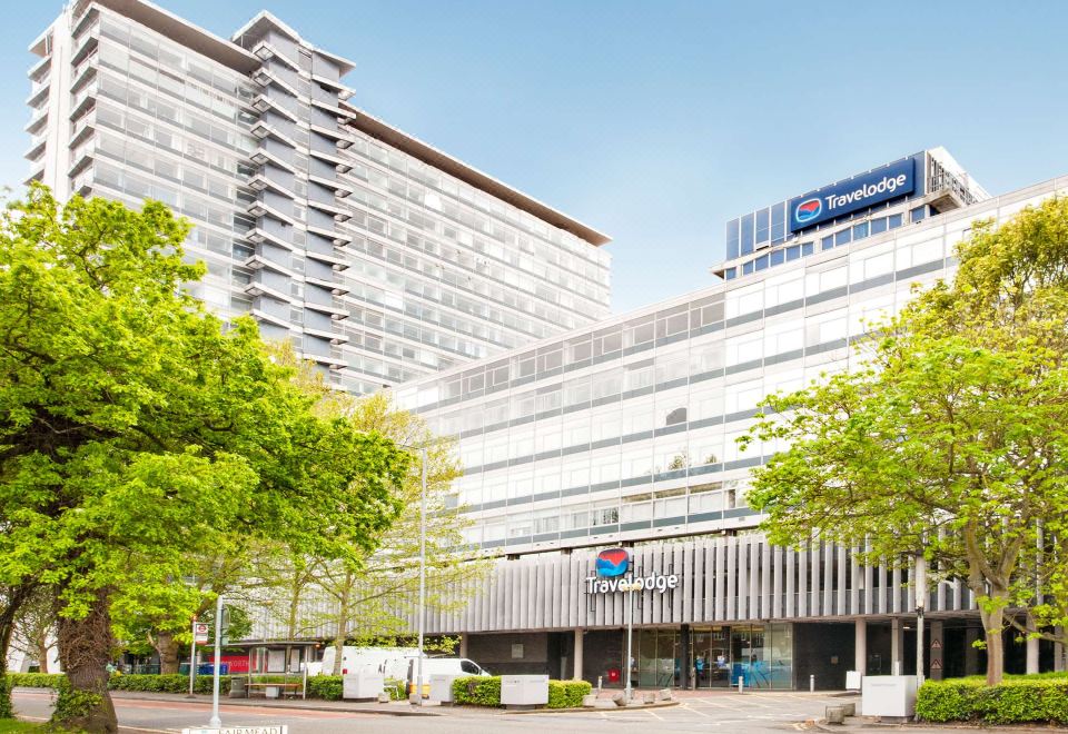 "a large , modern building with a red sign that reads "" bank "" in front of trees and bushes" at Travelodge London Chessington Tolworth