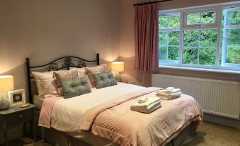 a well - arranged bed with pink bedding , two nightstands , and a lamp , next to a window with curtains and a view of trees at Leafy Suburban Bed and Breakfast