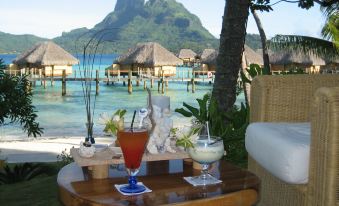 a table with two drinks and a view of the ocean in front of tropical houses at Le Bora Bora by Pearl Resorts