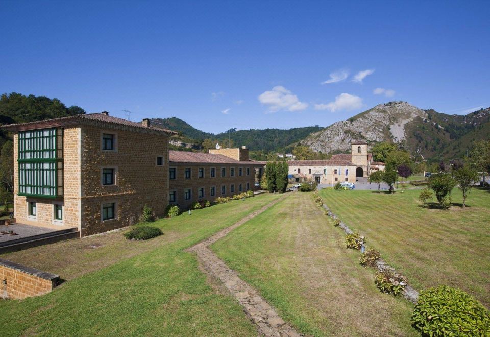 a large stone building surrounded by a grassy field , with mountains in the background , under a clear blue sky at Parador de Cangas de Onis