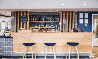 a bar with wooden counter and stools , blue stools in front of the counter , and shelves filled with various bottles at Holiday Inn Express Stockport