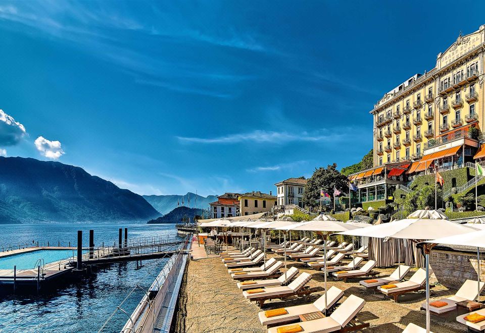 a large , white building with many umbrellas and chairs on the waterfront , under a clear blue sky at Grand Hotel Tremezzo