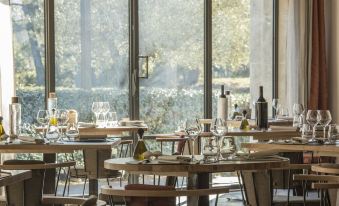 a restaurant with wooden tables and chairs , wine glasses , and bottles , set against a backdrop of large windows at Domaine la Pierre Blanche