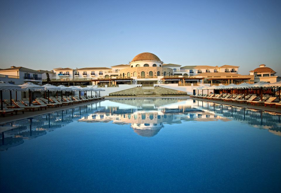 a large swimming pool surrounded by lounge chairs and umbrellas , with a building in the background at Mitsis Selection Laguna