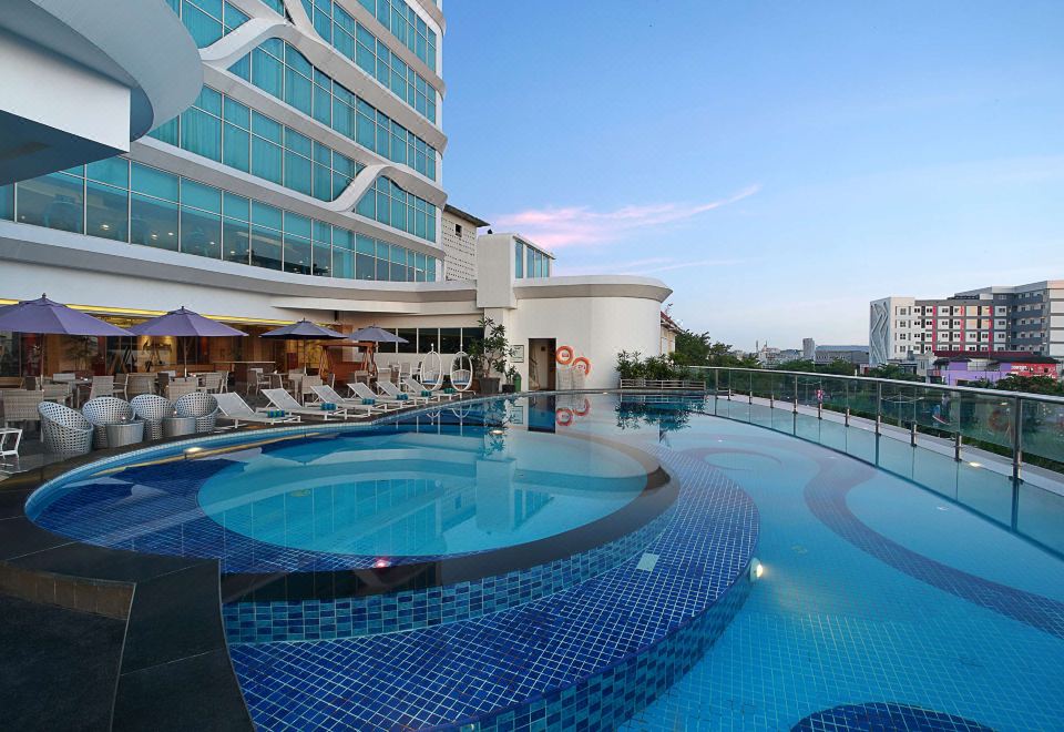 a large outdoor pool surrounded by a hotel , with several lounge chairs and umbrellas placed around the pool area at Galaxy Hotel Banjarmasin