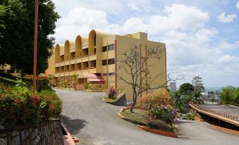 a large yellow building with a curved facade is surrounded by a stone wall and bushes at Ambassador Hotel