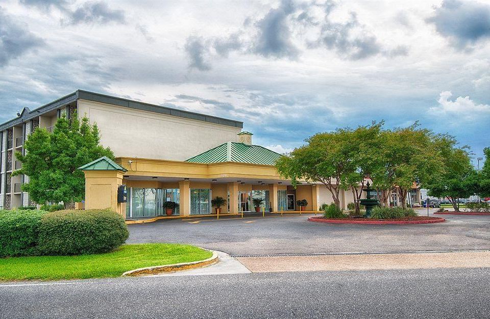 a large building with a green roof and yellow walls is surrounded by trees and bushes at Ramada by Wyndham Metairie New Orleans Airport