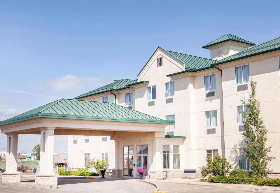 a holiday inn express hotel with its green roof and white walls , surrounded by trees and bushes at Ramada by Wyndham Estevan