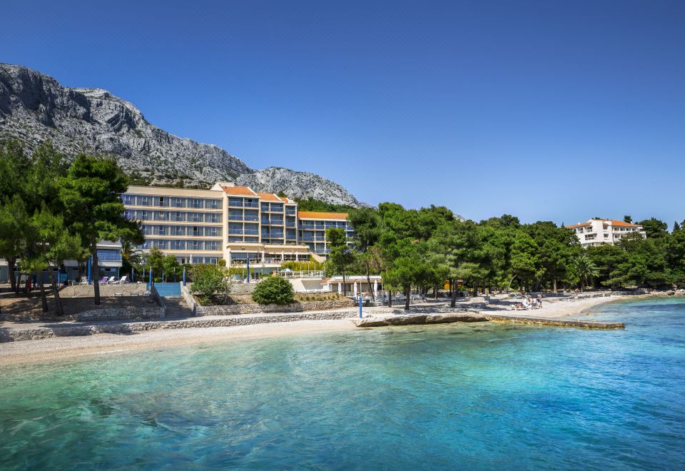 a large hotel on a beach with clear blue water and mountains in the background at Aminess Grand Azur Hotel