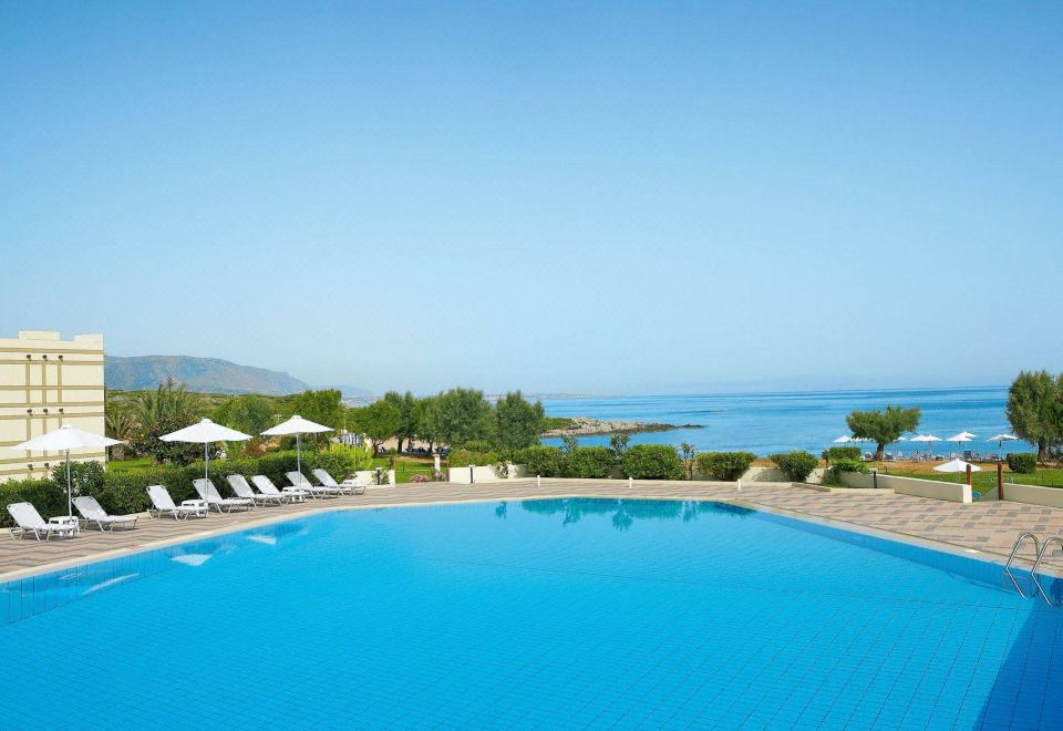 a large outdoor swimming pool surrounded by lounge chairs and umbrellas , with a view of the ocean in the background at Grecotel Meli Palace