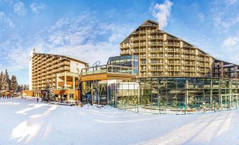 a modern hotel building with multiple floors , surrounded by snow - covered trees and a clear blue sky at Rila Hotel Borovets