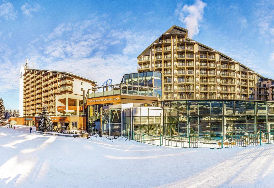 a modern hotel building with multiple floors , surrounded by snow - covered trees and a clear blue sky at Rila Hotel Borovets