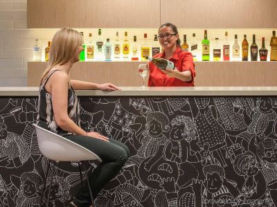 a woman is standing behind a counter in a bar , talking to a bartender about the drink at Ibis Melbourne Hotel and Apartments