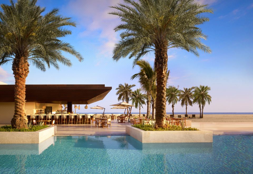 a beautiful outdoor pool area with palm trees and a beachfront restaurant , under a blue sky at Nobu Hotel Los Cabos