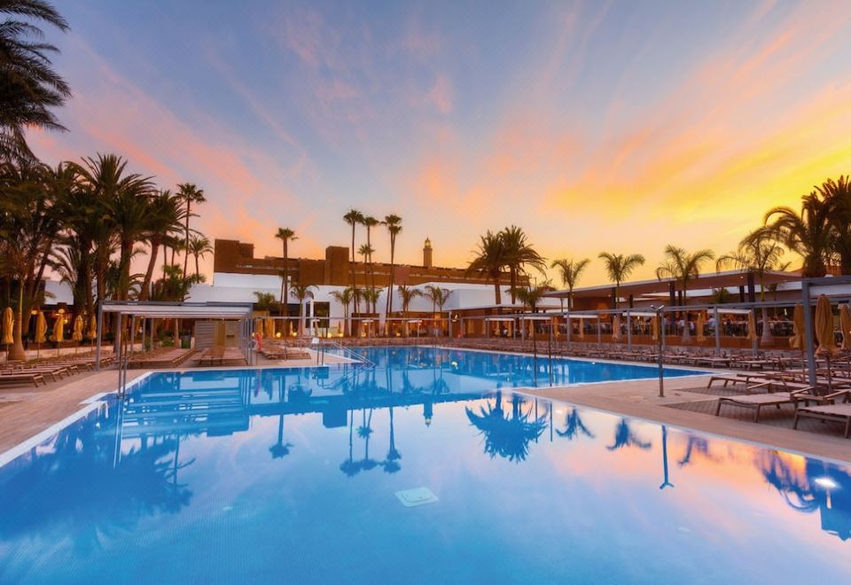 a large outdoor swimming pool surrounded by palm trees , with a beautiful sunset in the background at Hotel Riu Palace Oasis