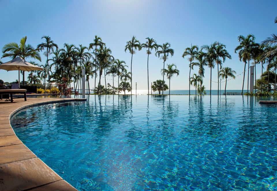 a large outdoor pool surrounded by palm trees , with the ocean visible in the background at Mindil Beach Casino Resort