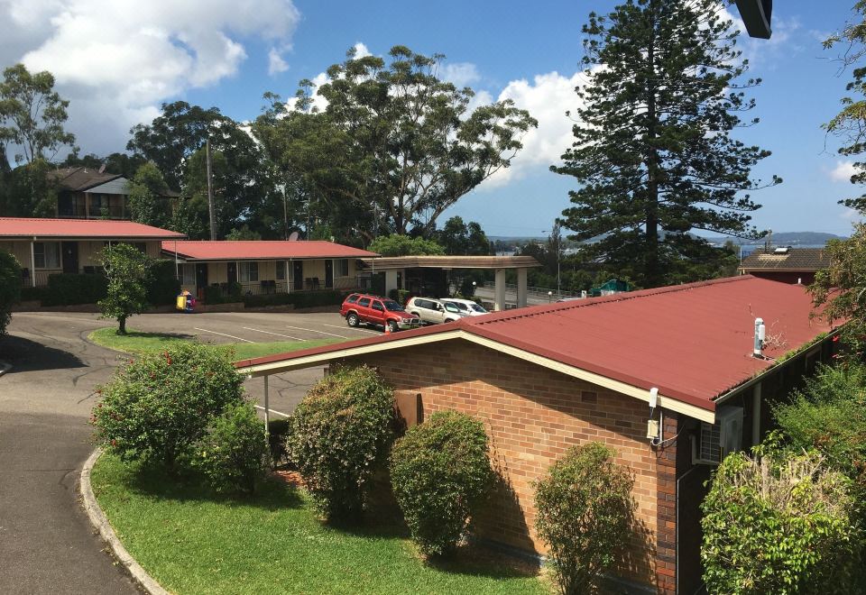 a view of a red - roofed building and parking lot with trees , cars , and a green lawn at Waterview Gosford Motor Inn