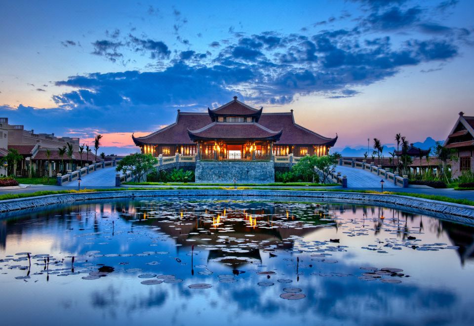 a traditional chinese building with a red roof and white walls is reflected in the water below , creating an eerie atmosphere at Emeralda Resort Ninh Binh