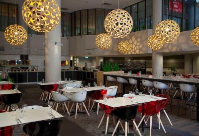 a modern restaurant with white tables and chairs , a bar area , and hanging lights in the background at Ibis Melbourne Hotel and Apartments