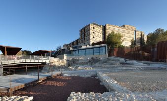 a modern building with a glass facade is situated on a cliff , overlooking a rocky area and water at Parador de Lorca