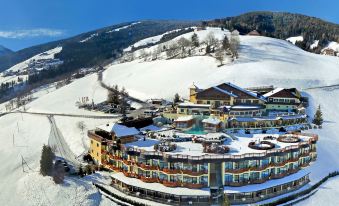 a snow - covered mountainous landscape with a large hotel complex at its base , under a clear blue sky at Alpin Panorama Hotel Hubertus